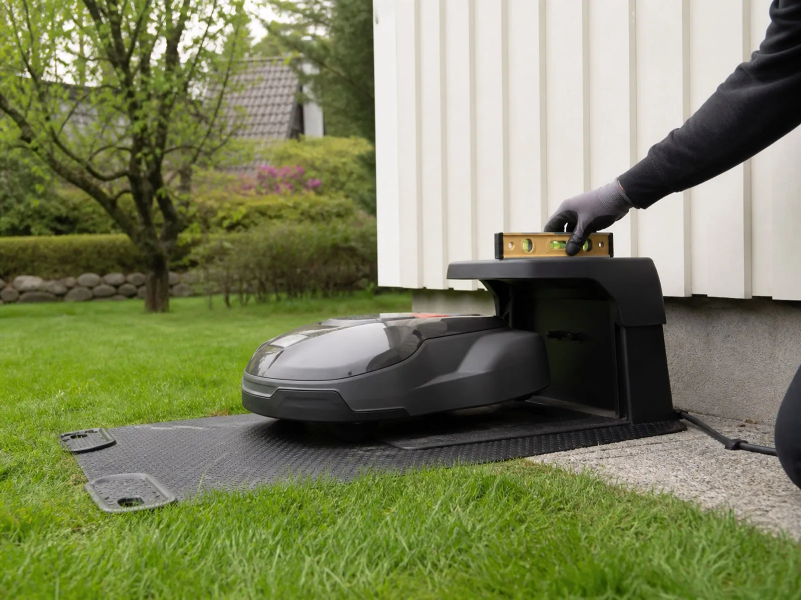 Laddstation för robotgräsklippare placerad på plan gräsyta nära husvägg.